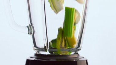Beyaz arka planda şeffaf blender kasesinin içinde dilimlenmiş çiğ sebzeler. Yeşil organik sebzeler yavaş çekimde cam karışımına dökülüyor. Vitamin kokteyli hazırlanıyor..