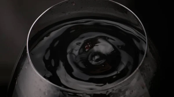 超遅い動きの閉鎖で透明なワイングラスを落下赤ワイン液滴 円形の上面に滑らかなアルコール飲料表面リップル 黒の背景にゴブレットの中に手を振るおいしいブドウ飲料 — ストック写真