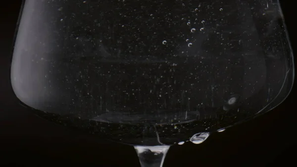 Small bubbles swirling inside full goblet of water in super slow motion close up. Crystal wineglass filled with clear liquid on black background. Drops of mineral drink falling from glass surface.