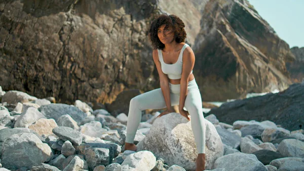 Young strong woman training yoga firefly pose on big stone rocky seacoast. African american focused girl making asana for training balance strength on nature. Sportswoman standing on hands outdoors.