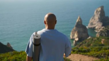 Yoga yapan adam uçurumun kenarındaki minderi tutuyor. Odaklanmış sporcu yaz güneşinde okyanus kıyısına bakıyor. Yetişkin turistler sakin deniz manzarasının tadını çıkarıyorlar. Sahildeki antrenman yerini seçen atletik bir adam.