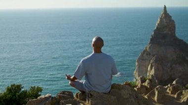 Yogacı adam altın güneş ışığında güzel bir uçurumun kenarında meditasyon yapıyor. Nilüfer pozisyonunda nefes alan tanınmamış atletik kel adam doğayla dengede duruyor. Güzel okyanusta huzurlu bir an.