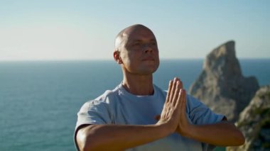 Yoga yapan bir sporcu okyanus manzarası. Odaklanmış bir adam güzel bir uçurumun kenarında namaste pozisyonunda meditasyon yapıyor. Kel yakışıklı adam güneş ışığında dışarıda dinleniyor. Farkındalık konsantrasyon konsepti.