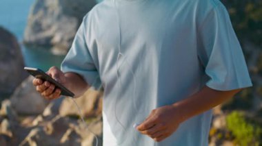 Okyanus kıyısındaki uçurumda akıllı telefon kullanan erkek elleri. Sporcu adam kulaklığı açık havada tutuyor. Tanınmayan kullanıcı sosyal medyayı kaydırıyor. Denizde podcast seçin. Akıllı teknoloji konsepti.