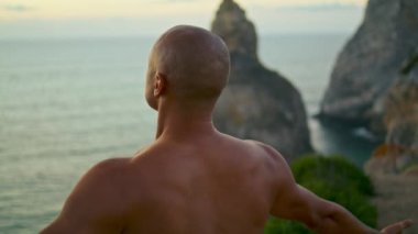 Okyanus kayalıklarında meditasyon yapan sakin bir yogi. Günbatımı deniz manzarasında kaslı bir atletik. Tanınmayan kel adam dışarıda yoga yapıyor. Üstsüz jest yapıyor. Güzel bir yerde egzersiz..