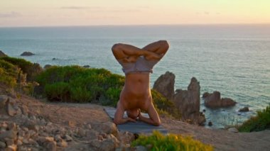 Kaslı bir adam, okyanus kayalıklarında günbatımı manzarasında yoga yapıyor. Kel sporcu, güneş ışığında üstsüz kafa egzersizi yapıyor. Yogi tren dengesi kayalık sahilde. Zen fitness konsepti gibi.