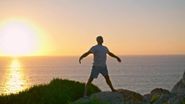 İnsan silueti akşam güneşli plajlarda yoga yapıyor. Güzel okyanus uçurumu manzarası. Tanınmayan adale atleti, deniz kayası üzerinde asana antrenmanı yapıyor. Yogi insanı dışarıda esniyor. Uyumlu yaşam tarzı kavramı