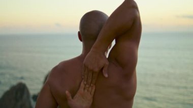 Güçlü yogiler, akşam okyanusa yakın çekimde manevi asana çalışırlar. Kaslı adam geri görüşü. Tanınmayan kel sporcu, güzel deniz kayalıklarında el kilidi açıyor. Doğa kavramı ile uyum bağlantısı.
