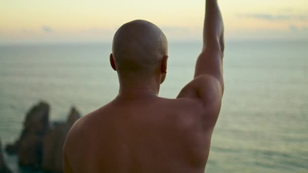 海の夕日の眺めで腕を伸ばして筋肉の男を閉じます 運動選手は屋外でトップレスロックを行う 未知のフィットヨギは海で精神的な練習を行う呼吸します 穏やかな夜のワークアウトのコンセプト — ストック動画