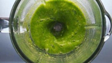 Üst manzara yeşil smoothie yüzeyi mikser kasesinde kıvrılıyor. Yakın çekim. Gri arka planda cam karıştırıcının içinde organik sebzeler var. Sebze vitamin kokteyli süper yavaş çekimde karışıyor..