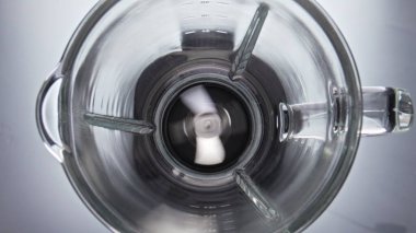 Üst görünüm boş elektrikli blender süper yavaş çekimde çalışıyor. Metal bıçaklar gri arka planda yemek yemeden cam kasede dönüyorlar. Yemekleri karıştırmak için yararlı modern mutfak gereci.