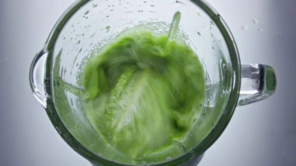 Closeup green vegetables mix in electric blender close up top view. Sliced veggies fruits herbs blending in glass bowl in super slow motion. Preparing vegan vitamin healthy smoothie in mixer.