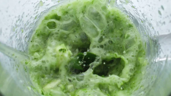 Greens mixing with vegetables in blender bowl close up. Fresh leafs kale parsley blending glass mixer in super slow motion. Green vitamin blend foaming swirling top view. Vegetarian nutrition concept.
