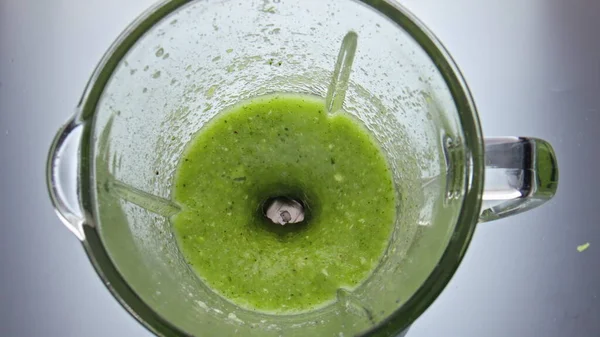 Green veggies cocktail mixing in electric blender close up top view. Tasty vitamin blend swirling in glass bowl super slow motion. Fresh raw vegetables fruits herbs blended for healthy smoothie.