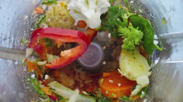 Mixed vegetables blending in blender bowl super slow motion close up. Mixer blades cutting sliced veggies for vitamin cocktail top view. Smoothie from broccoli pepper cauliflower potato salad carrot.