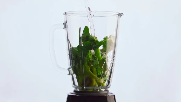 Fresh vegetables dropped into glass blender pouring with clear water to make delicious healthy smoothie close up. Liquid stream flowing at sliced green veggies in mixer bowl super slow motion.