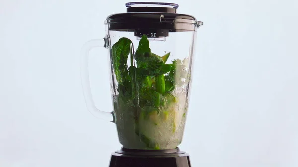 Electric blender preparing vegetable smoothie in super slow motion close up. Green organic veggies herbs fruits blending in glass mixer bowl white background. Cooking healthy vegetarian cocktail.