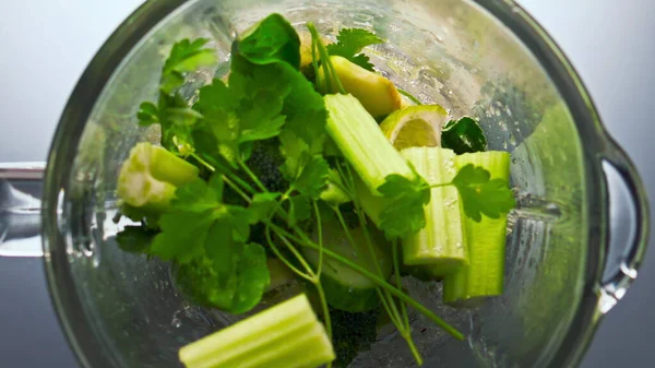 Pieces raw green vegetables inside blender preparing for vitamin cocktail in super slow motion closeup. Top view sliced veggies fruits herbs start blending in glass mixer bowl. Healthy vegetarian food