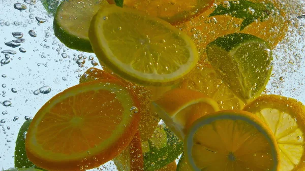 Pieces juicy citrus fruit dropped with mint in clear water for making tasty lemonade close up. Fresh sliced orange lemon lime floating with bubbles in super slow motion. Vitamin food in healthy life.