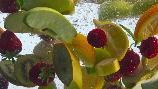 Mix sliced fresh fruits dropped clear water floating with bubbles in super slow motion close up. Various juice ingredients washing for preparing vitamin juice. Tasty banana orange kiwi strawberry mint