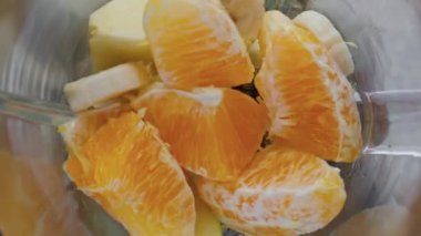 Dilimlenmiş sulu meyvelerle dolu elektrikli blender. Cam kasede karıştırılmış lezzetli muzlu elma mandalinası süper yavaş çekimde. Karıştırıcıda organik vejetaryen içeceği pişiriyorum..