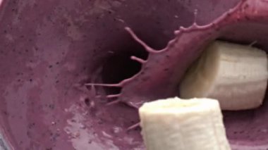 Muz düşen milkshake blender kasesinin üst görüntüsü. Süt böğürtlenli smoothie yakın plan hazırlıyor. Meyve suyu yavaş çekimde sıçrıyor. Konteynırda protein laktoz karışımı. Sağlıklı tatlı.