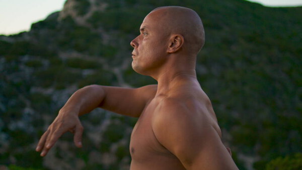 Fit yoga man concentrating at green landscape closeup. Focused bald athlete meditating doing spiritual practices topless in evening. Muscular guy perform namaste gesture relaxing workout in mountains