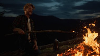 Joyful man camping outside on dark evening. Close up mountains traveler relax on nature alone. Adventurous millennial enjoy watch fire burning on campfire. Recreation freedom wanderlust youth concept.