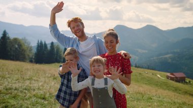 Laughing family waving hands on camera standing mountain hill. Happy parents with children posing on beautiful green slope sunny day. Cheerful couple with two kids enjoying holiday in summer nature.