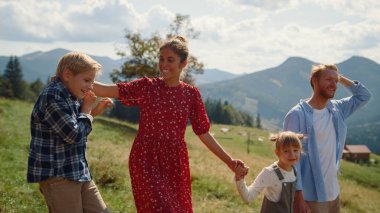 Cheerful parents smiling walking with children on green grass mountain valley. Carefree family enjoying summer vacation on beautiful nature. Cheerful couple with kids feeling happy together at holiday