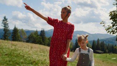 Walking woman relax with daughter on green meadow. Smiling mother holding girl hand on family stroll sunny day. Happy mom showing mountain views to cute child outdoors. Leisure together concept.