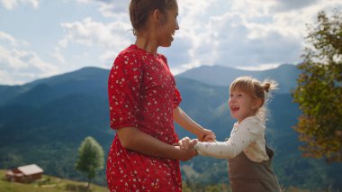 Side view of cheerful mother jumping with daughter dancing on mountain hill close up. Smiling woman with cute girl holding hands bouncing outdoor. Happy mom having fun with child at summer holiday.