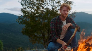 Active traveler camp forest in mountains. Close up redhead hiker sit bonfire on nature evening sunset. Cheerful camper throw log wood in fire. Young tourist spend weekend. Leisure relax travel concept