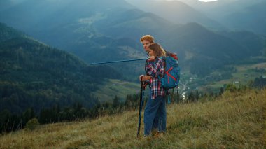 Sırt çantalı çiftler manzara dağ manzarasının tadını çıkarırlar. İki yürüyüşçü yeşil tepenin doğasında rahatlar. Sportif turistler güneşin doğuşuna bakarlar. Aktif yürüyüşçüler tatil maceralarında sırt çantası takarlar. Seyahat kavramı.