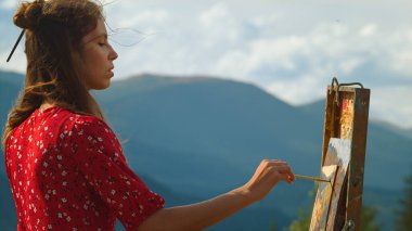 Yakın plan kadın ressam yaz dağlarında fırça darbesi yapıyor. Yetenekli kız doğa manzaralı resim sehpası. Profesyonel sanatçı doğadan resim çizer. Sanat eseri yapan genç ve ilham verici bir bayan.