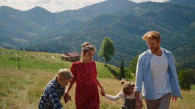 Positive family fooling on nature coming on top green hill. Joyful couple have fun with happy children outdoors sunny day. Cheerful parents walking with kids in beautiful mountains summer time.