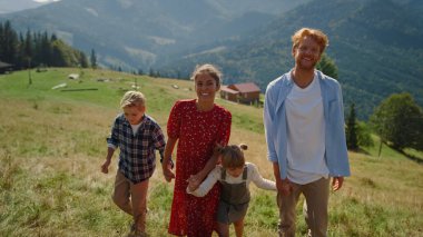 Playful parents have fun with children on grassy hill sunny day. Happy family fooling walking in beautiful mountains. Joyful couple with kids coming on top green slope summer weekend. Leisure concept.
