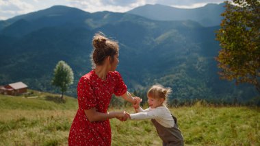Joyful woman jumping with daughter enjoying family leisure on green meadow. Young mother holding girl hands moving actively in mountains. Happy mom have fun with cute child summer vacation outdoors.
