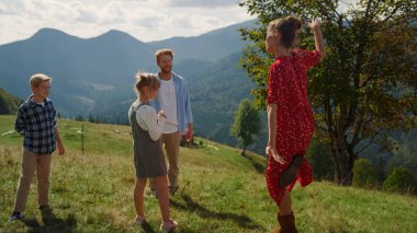 Young parents playing funny games with children on green mountain slope. Cheerful mother dancing showing moves to family. Joyful couple have active leisure with kids on beautiful nature summer day.