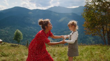 Happy mother enjoy funny dancing with daughter on green mountain hill. Cheerful woman teaching blonde girl dance element on nature. Pretty mom in red dress spending active summer holiday with child.