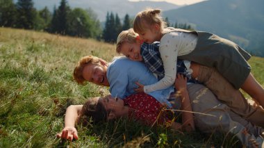 Playful family piled on each other lying green grass mountain hill close up. Happy parents playing with smiling children on summer nature. Joyful couple having fun with kids outdoors sunny day.