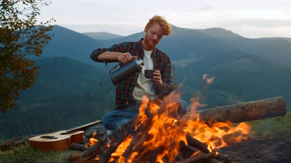 Relaxed tourist enjoy camping lifestyle by bonfire. Chill guy drink pour tea outside in mountains closeup. Young man hold thermos on hiking vacation. Redhead traveler spend evening. Adventure concept.