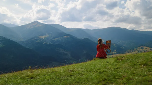 Back view of woman painter painting picture of gorgeous mountain landscape outdoors. Unknown girl drawing on easel sitting green hill summer holiday. Unrecognizable talented artist working on nature.