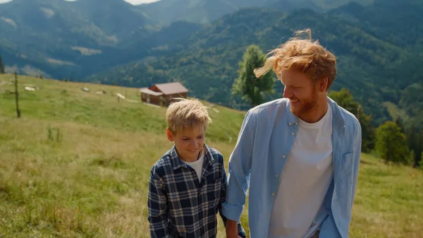 Ginger young man speaking with son on mountains walk closeup. Smiling father ruffling boy hair on green hill. Redhair parent with child have family stroll spending summer vacation on green nature.