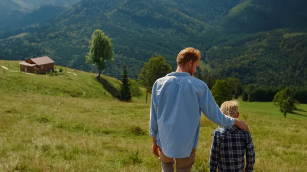 stock image Back view of red hair father with son walking on green hill sunny day. Young carefree parent hugging blond boy going down mountain slope. Unknown man spending summer vacation with child on nature.