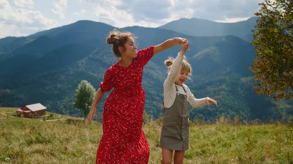 Cheerful mother dancing with daughter on green meadow. Happy woman holding hands with cute blonde girl relaxing on mountain hill sunny day. Positive mom with child moving bodies in dance summer time.