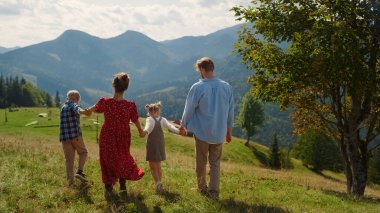 Back view of young family walking on mountains hill sunny day. Carefree trip together on green grass beautiful slope. Happy parents holding children hands enjoying summer vacation outdoors.