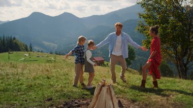Playful family having fun summer vacation in mountains. Happy parents enjoying active funny games on beautiful green hill. Joyful woman fooling touching children playing on grass with redhair man.