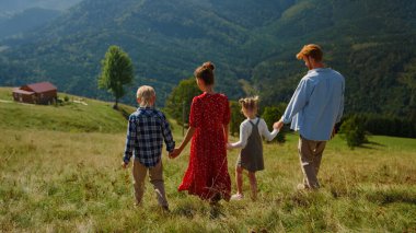 Back view of walking family going down green hill. Unknown parents enjoying summer vacation in mountains with children. Carefree couple with kids relaxing looking wonderful landscape summer nature.
