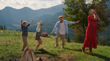Active family playing dancing funny moves on green grass beautiful hill. Joyful ginger father jumping with children near young mother closed eyes. Parents have fun with children on mountain slope.